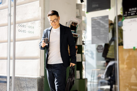 Asian businessman with coffee going from entrance of building. Smiling modern adult successful man...