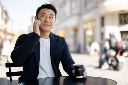 Asian businessman drinking coffee and talking on mobile phone at outdoor cafe. Concept of rest and leisure. Modern adult successful man wearing suit. Blurred background of sunny city streetの写真素材