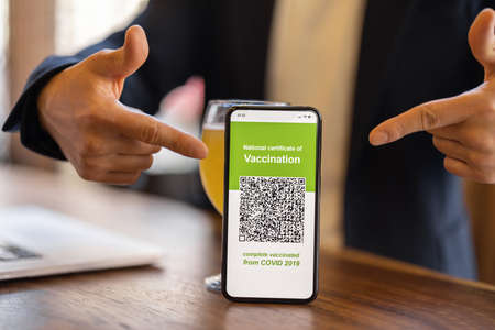 Cropped image of man showing smartphone with app of Coronavirus Vaccination in cafe. Concept of health protection during pandemic. Idea of rest and leisure. Wooden table with laptop and cocktailの写真素材