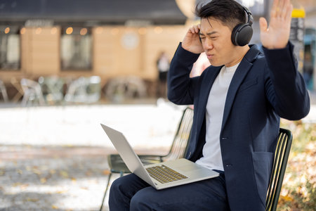 Irritated asian businessman in earphones working on laptop outdoors. Concept of remote work and freelance. Dissatisfied modern adult successful man wearing suit sitting on chair. Sunny dayの写真素材