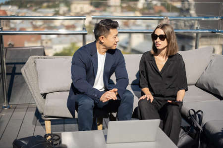 Businessman and businesswoman talking on balcony on building roof. Caucasian woman and asian man sitting on sofa at desk with laptop. Concept of business cooperation. Blurred background of cityの写真素材
