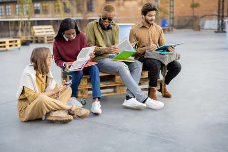 Multiracial students reading books while sitting on wooden bench outdoors at day. Concept of education and learning. Idea of student lifestyle. Young modern guys and girls at university campusの写真素材