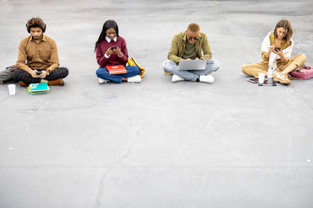 Multiracial students sitting and using gadgets on asphalt at university campus. Concept of education. Remote and e-learning. Idea of student lifestyle. Girls and guys wearing casual clothesの写真素材