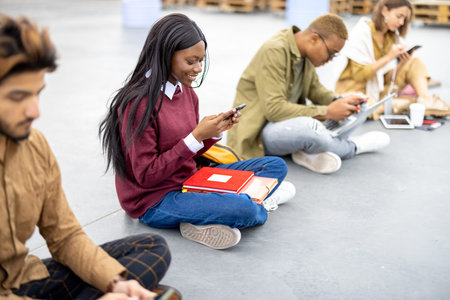 Row of multiracial students sitting and using smartphones on asphalt at university campus. Concept...
