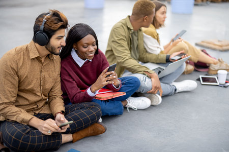 Multiracial students sitting and using digital devices on asphalt at university campus. Concept of...