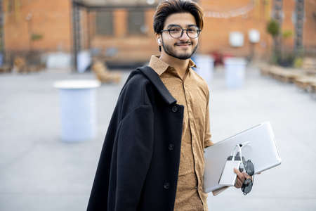 Young indian man with gadgets looking at camera outdoors. Handsome smiling stylish guy in glasses. Concept of education and learning. Idea of student lifestyle. Male person at university campusの写真素材