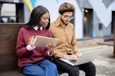 Black girl with digital tablet watching something on laptop with indian guy outdoors. Concept of remote and e-learning. Students lifestyle. Multiracial students sit on wood bench at university campusの写真素材