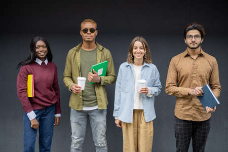 Multiracial students standing in row and looking at camera on dark and blue background. Concept of education and learning. Idea of students lifestyle. Young guys and girls holding notebooks and coffeeの写真素材
