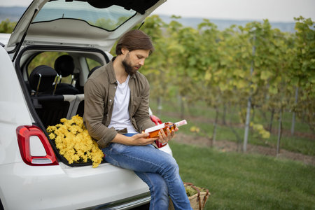Man holds bottle with local wine near vineyards, sitting at car trunk in countryside. Concept of...