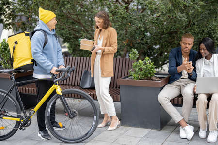 Courier in medical mask giving food order to business woman outdoors. Concept of shipping and logistics during Coronavirus pandemic. Idea of break on job. Smiling businessman and businesswomanの写真素材