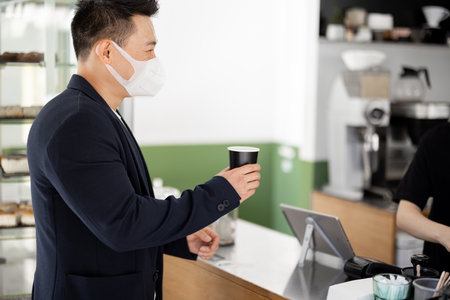 Asian businessman in medical mask with coffee going in store. Concept of health protection during Coronavirus pandemic. Modern adult successful male person wearing suitの写真素材