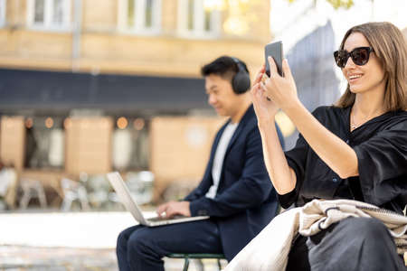 Two businesspersons sitting separately on chairs on the old city street outdoors. Woman taking photo on phone, man working o laptop. Concept of city life. Sunny autumn dayの写真素材