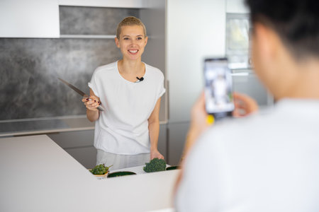 Man taking video on smartphone of caucasian woman cooking vegetarian salad on kitchen. Concept of video blogging. Idea of healthy and diet eating. Smiling girl with knife looking at mobile phoneの写真素材