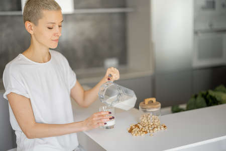 Young caucasian woman pouring vegetable milk from jug in glass at table on kitchen at home. Concept of healthy vegan life. Idea of domestic lifestyle. Beautiful slim focused girl. Interior of modern flatの写真素材