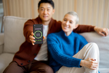Asian man sitting on sofa with his caucasian wife and showing smartphone with home air condition...
