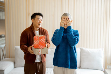 Asian man giving gifts to his girlfriend with closed eyes. Concept of holiday, event and birthday. Idea of family relationship. Happy couple in casual clothes. Interior of living room in modern flatの写真素材