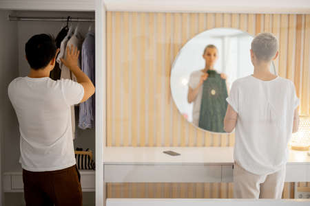 Asian man choosing clothes in wardrobe while his caucasian wife looking dress on herself in mirror. Couple getting ready to spend time together in public place. Family relationship. Modern apartmentの写真素材