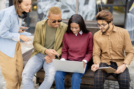 Multiracial students watching something on laptop while sitting on wooden bench outdoors. Concept of...