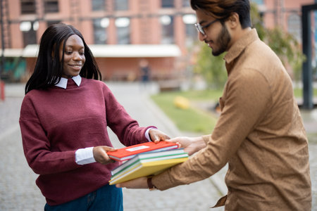 Indian man giving books to black girl outdoors. Concept of education and learning. Idea of students lifestyle. Smiling friends at university campusの写真素材