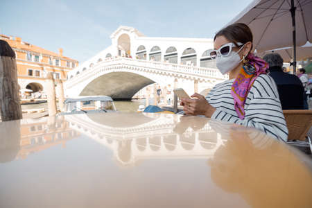Young woman in medical mask sitting with phone at outdoor cafe near Grand Canal and Rialto bridge in Venice. Concept of social rules for travelers during pandemic. Idea of a happy vacation in Italyの写真素材