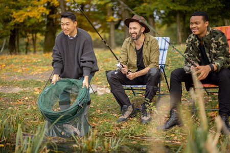 Pleased multiracial male friends resting and fishing on river or lake coast. Concept of leisure,...