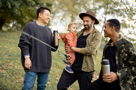 Smiling multiracial male friends resting in nature at autumn. Concept of leisure, weekend and entertaiment. Idea of friendship and spend time together. Caucasian man with fishing rod and little sonの写真素材