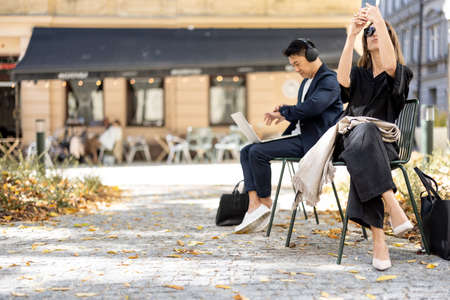 Two businesspersons sitting separately on chairs on the old city street outdoors. Woman taking photo on phone, man working o laptop. Concept of city life. Sunny autumn dayの写真素材