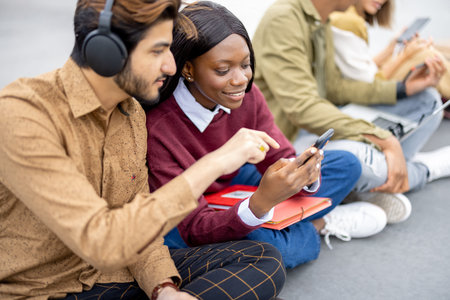 Multiracial students sitting and using digital devices on asphalt at university campus. Concept of...