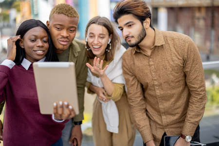 Multiracial students watching something on digital tablet outdoors. Concept of education. Remote and e-learning. Idea of student lifestyle. Smiling young friends at university campusの写真素材