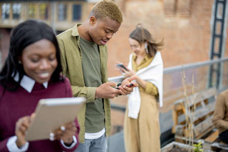 Focused multiracial students using digital devices on balcony at university campus. Concept of education. Remote and e-learning. Idea of student lifestyle. Persons wearing casual clothesの写真素材