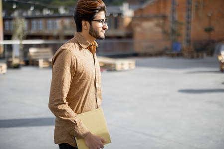 Young indian man with gadgets looking at camera outdoors. Handsome smiling stylish guy in glasses. Concept of education and learning. Idea of student lifestyle. Male person at university campusの写真素材