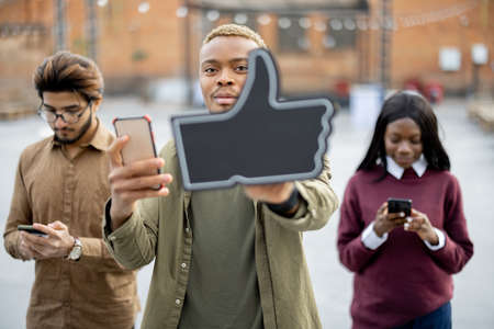 Young black guy holding smartphone and showing thumb up gesture on background of his multiracial friends with mobile phones at university campus. Concept of education and learning. Student lifestyleの写真素材