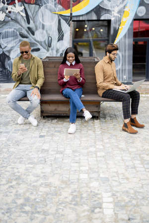 Multiracial students using gadgets outdoors. Concept of education. Remote and e-learning. Idea of students lifestyle. Young guys and girl sitting on wooden bench at university campusの写真素材