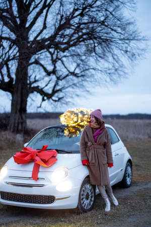 Woman in front of her white car with illuminated Christmas tree and festive red bow outdoors. Concept of New Year preparation. Idea of a fairy tale and a festive mood in wintertimeの写真素材