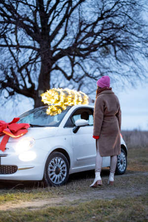 Woman walks in front of her white car with illuminated Christmas tree and festive red bow outdoors. Concept of New Year preparation. Idea of a fairy tale and a festive mood in wintertimeの写真素材