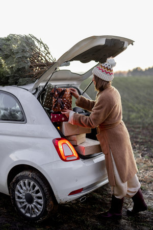 Woman packing gifts into the car with Christmas tree on a rooftop on nature at dusk. Getting ready for a New Year holidays. Idea of a Christmas mood. Woman wearing fur coat and hatの写真素材