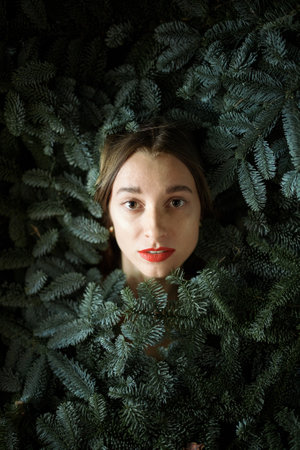 Face portrait of a young woman covered with branches of nobilis. Concept of female beauty and freshness. Coniferous branches and nature for facial care. Idea of winter holidaysの写真素材