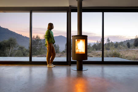Woman enjoy great view on mountains while sitting near fireplace at modern living room at sunset . Concept of rest in houses or cabins on nature. Solitude in nature and escape from everyday lifeの写真素材