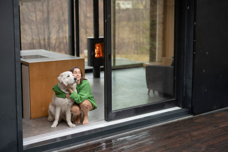 Young woman with her dog at modern house on nature. Girl in green sweater standing near the window and look outside. Concept of comfort and happy modern lifeの写真素材