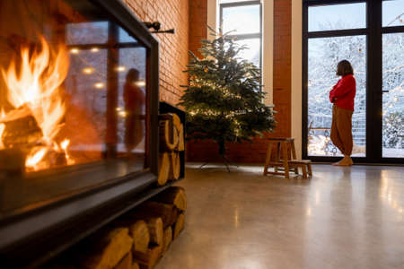 Cozy living room interior with Christmas tree and burning fireplace. Woman in red sweater near the window. Concept of home comfort in winter. Idea of loneliness and celebration aloneの写真素材