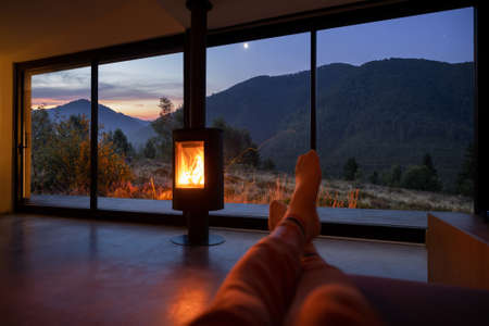 Woman resting near fireplace at modern house with great view on mountains at sunset. Womens legs on background of landscapeの写真素材