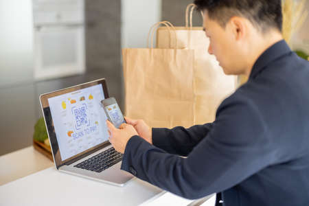 Man using smartphone for payment in online store on laptop on kitchen at home. Concept of online shopping and e-commerce. Idea of healthy and vegetarian eating. Person sit at table with shopping bagsの写真素材