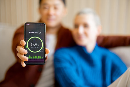 Asian man sitting on sofa with his caucasian wife and showing smartphone with home air condition app. Concept of smart home. Idea of domestic lifestyle. Smiling multiracial couple in modern apartmentの写真素材