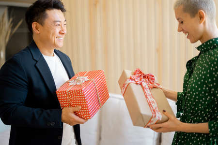 Asian man and young caucasian woman giving gifts each other. Concept of holiday, event and birthday. Idea of family relationship. Happy smiling couple sitting on sofa. Interior of modern apartmentの写真素材