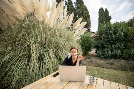Young caucasian woman having video call on laptop computer while sitting at wooden table on green mediterranean lawn. Concept of weekend, rest and remote work on natureの写真素材