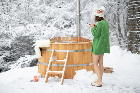 Woman taking photo of a wooden bath, during winter vacations at snowy mountains. Scandinavian lifestyle and and hot water treatments concept. Girl wearing sweater and hatの写真素材