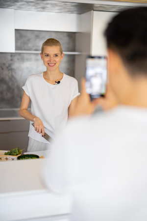 Man taking video on smartphone of caucasian woman cooking vegetarian salad on kitchen. Concept of...