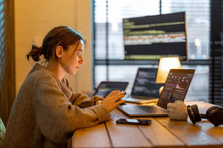 Young woman using phone while sitting at workplace with laptop and desktop computers at cozy home...