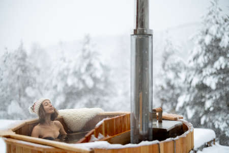 Woman relaxing in hot bath outdoors, enjoying thermal spa at snowy mountains. Winter holidays in the mountains, hot water treatments concept. Caucasian woman wearing winter hatの写真素材