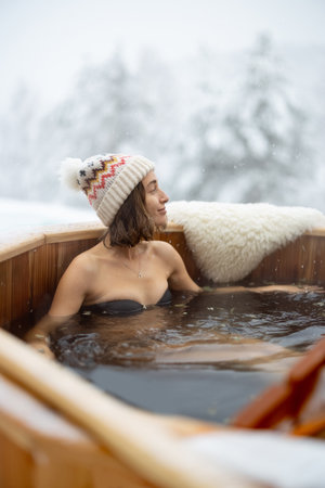 Woman relaxing in hot bath outdoors, enjoying thermal spa at snowy mountains. Winter holidays in the mountains, hot water treatments concept. Caucasian woman wearing winter hatの写真素材